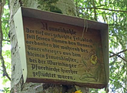 Tafel bei der Nonnenhöhle bei Wurmlingen mit Hinweis auf die Namensgebung Blick auf eine kleine überdachte Holztafel. Die Tafel ist an einem Baumstamm befestigt und erzählt, wie die Nonnenhöhle zu ihrem Namen kam.
