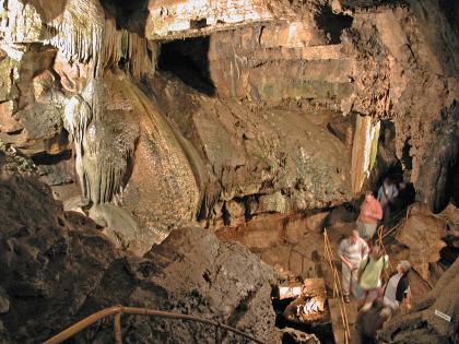 Besucher in der Erdmannshöhle bei Hasel Leicht abwärts gerichteter Blick in das Innere einer Höhle mit verschachtelten Wänden und Tropfsteinbildungen links und rechts. Rechts unten steigen Besucher eine Treppe zu einem Steg hinab.