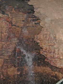 Das Foto zeigt einen Wasserfall in einer Höhle. Der Wasserfall befindet sich vor einer Höhlenwand, zwischen zwei vorstehenden Seitenwänden links und rechts.