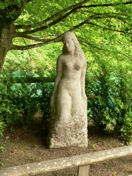 Skulptur der „schönen Lau“ am Blautopf Auf dem Bild sieht man die aus einem hellen Steinblock modellierte Skulptur einer Frau. Die Skulptur steht im Schatten eines Baumes und ist von Sträuchern umgeben.