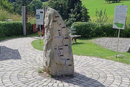 Stele und Infotafeln am Zugang zur Bregquelle bei Furtwangen Blick auf einen sich nach oben verjüngenden Steinblock. Auf einer Seite ist der Lauf der Donau eingraviert. Die Stele steht auf einem gepflasterten Weg. Im Hintergrund sind Hinweistafeln aufgestellt.