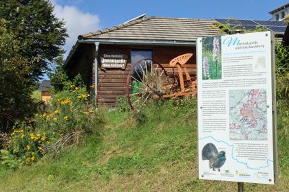 Infotafel auf dem Weg zur Bregquelle nordwestlich von Furtwangen Blick auf eine rechts im Bild stehende, mehrfarbige Hinweistafel auf dem Weg zum Naturdenkmal Donauquelle. Links, neben der Tafel, steht ein Holzhaus an einem Wiesenhang.