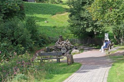 Auf dem Weg zur Bregquelle nordwestlich von Furtwangen Das Bild zeigt einen zum Hintergrund hin abfallenden Weg mit Sitzbänken und Bäumen, der zu einer Quelle führt. Blickpunkt der Quelle ist eine große Steinfigur. Dahinter steigt ein Wiesenhang auf.