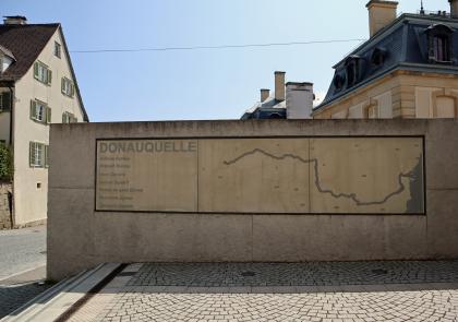Schautafel mit dem Flusslauf der Donau auf dem Weg zur Donauquelle an der Schlossmauer in Donaueschingen Das Bild zeigt eine lange Schautafel, die an einer halbhohen Mauer angebracht ist. Auf der Tafel ist der Flusslauf der Donau von der Quelle bis zur Mündung aufgezichnet.