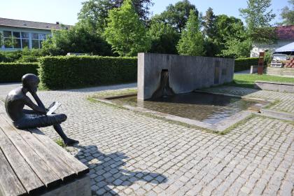 Die neue Neckarquelle im Stadtpark an der Möglingshöhe in Schwenningen Blick auf die neu gestaltete Quellfassung der Neckarquelle in einem Park. Die Quelle fließt aus einer langen, halbhohen Mauer in ein flaches viereckiges Becken und von dort weiter in schmale Rinnen. Eine lesende Figur sitzt links davon auf einer Bank.