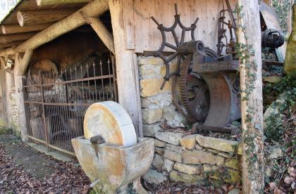 Im Freilichtmuseum „Historische Steinbrüche Pfaffenweiler” Seitlicher Blick auf einen teils aus Mauerwerk gebauten, teils hölzernen Schuppen. Der überdachte Schuppen ist zur linken Seite hin vergittert. Davor steht ein alter Wetzstein. Rechts an der Wand ist ein metallenes Zahnrad mit Drehkurbel angelehnt.