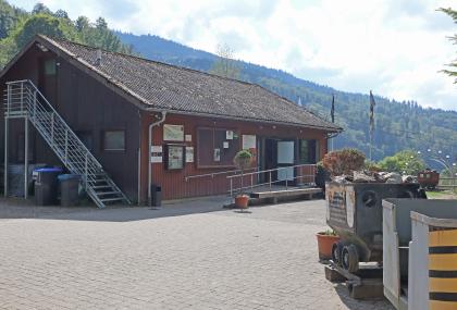 Grubenhütte des Silberbergwerks Teufelsgrund bei Münstertal Das Bild zeigt ein langes Gebäude aus braunem Holz mit seitlichem Eingang, das vor einer Kulisse mit bewaldeten Bergen steht. Das Gebäude gehört zum Besucherbergwerk Teufelsgrund. Rechts, auf dem gepflasterten Vorhof, sind alte Förderwagen abgestellt.