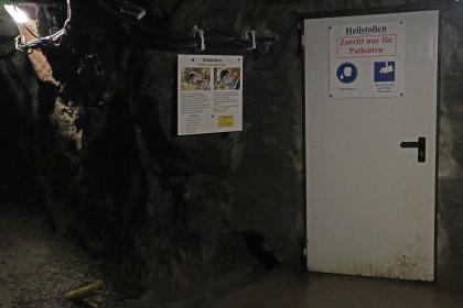 Eingang zum Asthmastollen im Silberbergwerk Teufelsgrund bei Münstertal Blick in einen dunklen Stollen des Besucherbergwerks Teufelsgrund. Eine Metalltür rechts sowie eine kleine Schautafel links davon weisen auf einen Heilstollen für Asthmakranke hin.