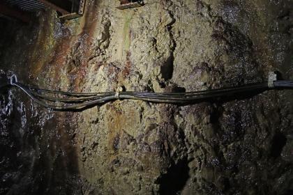 Von Wasser überrieselter Fels mit Sinterüberzügen im Silberbergwerk Teufelsgrund bei Münstertal Teilansicht einer grünlichen bis bräunlichen, an den Rändern schwarzgrauen Gesteinswand. Winzige Lichtpunkte reflektieren rieselndes Wasser auf dem Gestein. In horizontaler Richtung sind Kabelstränge an der Wand befestigt.