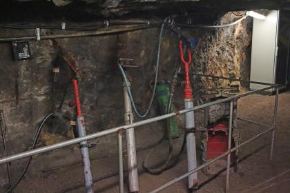 Druckluftbohrmaschinen im Silberbergwerk Teufelsgrund bei Münstertal Das Bild zeigt mehrere historische Bohrmaschinen im Besucherbergwerk Teufelsgrund. Die Bohrgeräte sind mit Luftschläuchen verbunden und stehen hinter einem Schutzgitter an einer Stollenwand.