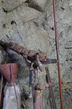 Historischer Pressluftbohrer und Bohrstange im Besucherbergwerk Finstergrund bei Wieden Das Bild zeigt einen alten, rostbraunen Pressluftbohrer, links angesetzt an einer grauen Gesteinswand. An die Wand gelehnt ist rechts noch eine lange dünne Bohrstange.