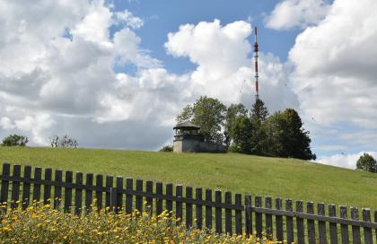 Im Mittelpunkt des Bildes steht ein kleiner gemauerter Pavillon mit Überdachung. Der Aussichtspunkt ist auf einer begrünten, flachen Bergkuppe. Ein paar Bäume und ein Sendemast stehen rechts vom Pavillon. Im Vordergrund verläuft ein Holzzaun.