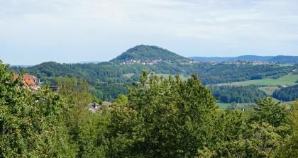 Blick vom Hohenstaufen zum Rechberg – Bei beiden Bergkegeln handelt es sich um in tektonischer Tieflage erhalten gebliebene Zeugenberge, die heute in 4–8 km Entfernung vor dem Trauf der Schwäbischen Alb liegen. Blick über Baumwipfel auf einen teils bewaldeten Höhenrücken. In der Bildmitte erhebt sich zudem ein bewaldeter Bergkegel. Am Fuß des Kegels sowie auf dem rechten Höhenrücken sind Häuser zu erkennen.