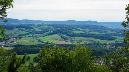 Blick vom Hohenstaufen über das Rehgebirge nach Südosten zum westlichen Albuch – Rechts im Hintergrund ist am Albtrauf der Talausgang des Filstals zu sehen. Weiter Blick über Baumkronen auf eine hügelige Landschaft mit Wiesen, Äckern und zahlreichen Waldflächen. Im Hintergrund verlaufen langgestreckte, flache Höhenzüge, die nach rechts hin abfallen.