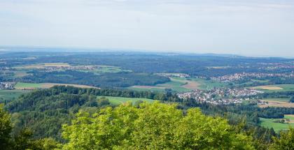 Über die vorgelagerte Stufenfläche im Niveau der Eisensandstein-Formation und die darunter anschließenden zertalten Unterjura-Flächen geht der Blick vom Hohenstaufen nach Westen zum Schurwald. Über Baumwipfel und einen vorgelagerten Hügelrücken geht der Blick auf eine wellige Landschaft mit mehreren Ortschaften und weiten Waldflächen.