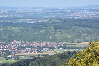 Blick vom Metzinger Rossberg über das Ermstal zum Hofbühl bei Metzingen-Neuhausen Blick aus großer Höhe auf eine hügelige Landschaft mit Feldern, Waldstücken und zahlreichen Ortschaften. Im Mittelgrund erhebt sich ein länglicher Hügelberg mit bewaldetem Rücken und terrassierten Hängen.
