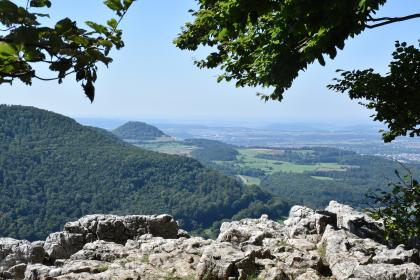In südwestlicher Richtung sieht man vom Metzinger Rossberg aus die Achalm und die darunter anschließende Stufenfläche im Mitteljura. Blick von erhöhtem Standort über ein Felsplateau im Vordergrund. Zum Hintergrund hin sieht man links einen bewaldeten Bergrücken sowie dahinter und rechts flache, bewaldete Hügel mit einem Bergkegel links.