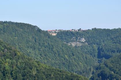 Blick vom Hanner Felsen nach Hülben, wo unter der Traufkante Aufschlüsse in der Untere-Felsenkalk-Formation (Oberjura) zu sehen sind Blick auf mehrere bewaldete Bergrücken. Der am entferntesten befindliche Berg weist eine waagerecht verlaufende Kuppe mit teilweiser Bebauung auf. Etwas darunter, rechts der Bildmitte, ist ein Gesteinsaufschluss am Hang erkennbar.
