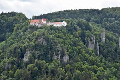 Blick auf einen bewaldeten Bergkegel. An den Seiten des Berges treten mehrere Felszacken hervor. Auf der flachen Kuppe des Berges stehen mehrere Gebäude, die miteinander verbunden sind.