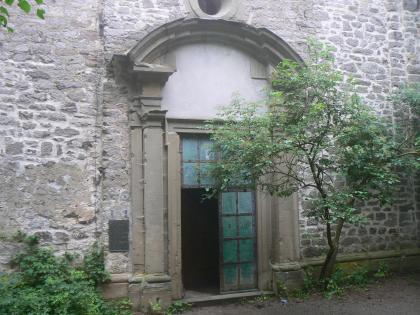 Ehemaliges Portal der Wallfahrtskirche auf dem Einkorn Blick auf ein steinernes, mit rundem Steindach versehenes Eingangsportal. Das Portal befindet sich in einer Fassade aus Mauersteinen. Die linke Hälfte der geteilten Eingangstür steht offen. Rechts daneben wächst ein kleiner Baum.