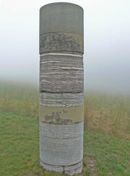 Geo-Säule auf dem Einkorn Blick auf eine hohe runde Steinsäule. Die Säule besteht aus mehreren Scheiben von unterschiedlichem Gestein, das aufeinandergestapelt ist. Die Farben wechseln dabei von grau zu senfgelb sowie rötlichen Wellenmustern.