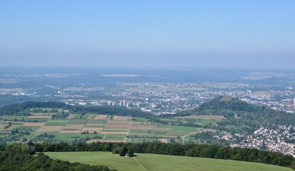 Der Vulkanschlot Georgenberg (rechts) bei Reutlingen und die links davon anschließende Stufenfläche des Mitteljuras (Wedelsandstein-Formation) Blick aus großer Höhe auf eine weite, dicht besiedelte Landschaft. Im Vordergrund erhebt sich eine Geländestufe mit bewaldeten Rändern sowie flachen Wiesen und Feldern. Rechts außen schließt sich ein kleiner Kegelberg der Stufe an.