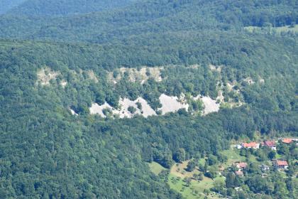 Felsen am Urselberg bei Lichtenstein-Unterhausen mit unterhalb anschließender Schutthalde Vergrößerter Blick auf einen langgestreckten bewaldeten Berghang, an dessen sichtbarer Flanke eine Felsreihe mit unterhalb anschließender Schutthalde erkennbar ist. Rechts unten, am Fuß des Berges, stehen Siedlungshäuser.