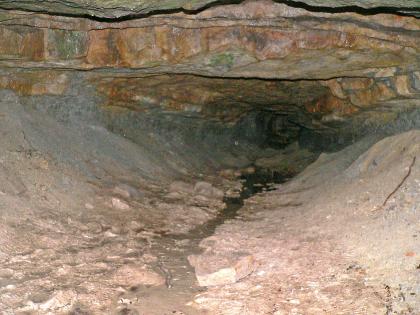 Blick in den Silberstollen südlich von Spiegelberg-Neulautern Das Foto zeigt das Innere eines Stollens. Zu sehen sind wannenförmige, graue Wände, eine rötlich braune, tiefhängende Decke sowie das trichterförmige Ende des Stollens.