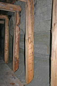 Das Bild zeigt eine graue Stollenwand in einem alten Bergwerk. Die Stollenwand wird durch mehrere Holzbalken gestützt. Oben links ist die durch Querbalken gestützte Stollendecke zu erkennen.