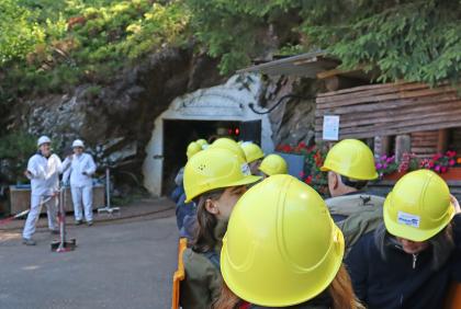 Das Bild zeigt eine Besuchergruppe des ehemaligen Bergwerks Finstergrund mit gelben Schutzhelmen. Der Eingang zum Bergwerk liegt in der Mitte des Bildhintergrunds.