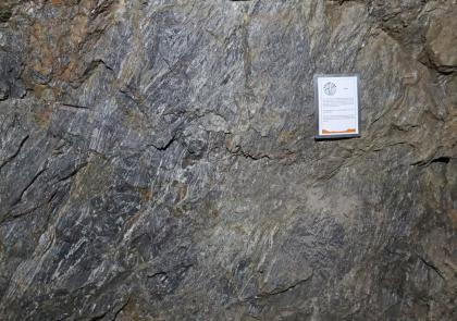 Geopunkt „Gneis“ im Besucherbergwerk Finstergrund bei Wieden Teilansicht einer alten Abbauwand in einem Besucherbergwerk; Farbe blaugrau bis graubraun, mit einer kleinen Infotafel rechts oben.