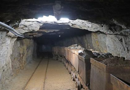 Besucherbergwerk Finstergrund bei Wieden – beladene Förderwagen der Grubenbahn stehen bereit für den Abtransport Blick auf einen breiten, niedrigen Tunnel mit zwei Spurbahnen für Förderwagen im Besucherbergwerk Finstergrund. Die nur im Vordergrund von einer Lampe beleuchteten Wagen auf der Spur rechts sind mit Abbaumaterial beladen. Die linke Spur ist frei.