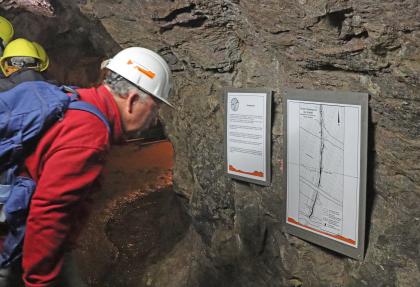 Besucherbergwerk Finstergrund bei Wieden – Schautafel zur Geologie des Finstergrundganges Seitlicher Blick auf zwei kleine Schautafeln, die an der dunkelgrauen Wand eines Besucherbergwerks angebracht sind. Ein Besucher mit weißem Schutzhelm schaut sich die Tafeln an.