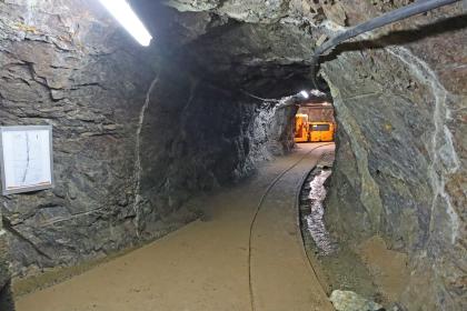 Besucherbergwerk Finstergrund bei Wieden – Querschlag zwischen dem Stollen 5 mit der Haltestelle der Grubenbahn und dem Wernerstollen Blick in den tunnelartigen Durchgang eines Bergwerkstollens. Am Ende des Stollens, rechts der Bildmitte, ist ein beleuchteter Querstollen erkennbar.