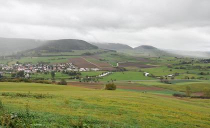 Blick vom Wartenberg über die Donauaue zum Kapf bei Gutmadingen und rechts hinten zum Fürstenberg Das Foto zeigt eine meist grüne, hügelige Landschaft. An den Hängen von im Hintergrund sich erhebenden Bergen sind auch Äcker erkennbar. Die Bergkuppen sind bewaldet. Im Mittelgrund schlängelt sich ein Fluss. Links hinten liegt eine Siedlung.