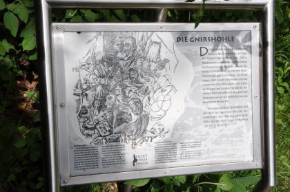 Informationstafel an der Gnirshöhle im Eiszeitpark bei Engen In einen Metallrahmen eingelassene Schautafel, die mit Wort und Bild von der Gnirshöhle während der Eiszeit erzählt. Im Hintergrund dichtes Blattwerk.