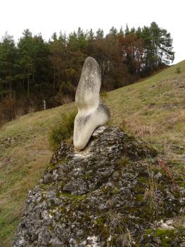 Am Fuß eines steilen, nach rechts ansteigenden und bewaldeten Hanges ist ein runder Felsenhügel zu sehen. Auf der Spitze dieses Hügels thront eine Statue aus weißlichem Material. Sie erinnert an eine sitzende Figur. 