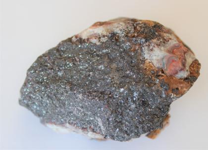 Hämatit aus der Grube Krunkelbach Das Bild zeigt ein kleines Gesteinsstück, das vergrößert fotografiert ist. Die vordere Seite des Handstückes ist grau, dahinter sind weißliche, rötliche und braune Farben zu erkennen.