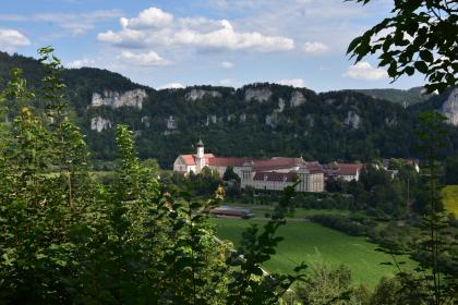 Blick auf das Kloster Beuron und das Donautal Blick von erhöhtem Standort auf eine große Klosteranlage mit hellen Mauern und roten Dächern. Vor dem Kloster breitet sich eine Wiese aus, hinter der Anlage steigen von Wald umschlossene weißgraue Felsspitzen auf.