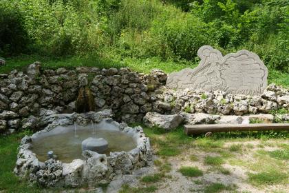 Schäferquelle im Grauental Blick auf eine runde, aus Felsgestein und Beton gefertigte Quellfassung. Der Quelltopf befindet sich links im Bild, vor einer aus Felsblöcken bestehenden Mauer. Rechts davon ist eine Steintafel zu sehen, mit dem Motiv eines Schäfers.