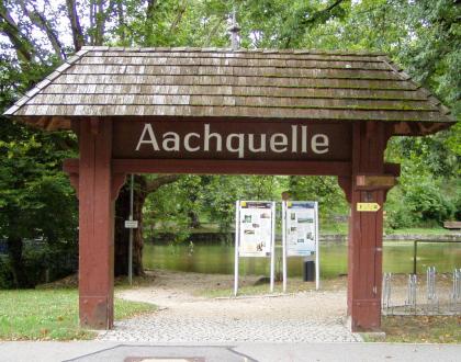 Die Aachquelle ist die am stärksten schüttende Karstquelle Deutschlands Blick auf ein großes, aus Holzpfosten gebautes und überdachtes Tor, das zur Aachquelle führt. Im Hintergrund sind ein Teil des Quellsees, das angrenzende belaubte Ufer sowie Hinweistafeln zu erkennen.