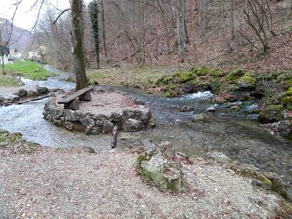 Von einer Ortschaft links hinten strömt ein Wasserlauf nach rechts am Betrachter vorbei. Links der Bildmitte umfließt er dabei eine kleine, von Felsgestein umrandete Insel mit Baum und Sitzbank. Das vordere Ufer ist flach, das hintere bildet ein Waldhang.