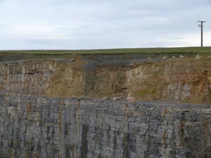 Großflächige, mittlerweile verfüllte Dolinenstruktur in einem Steinbruch bei Ditzingen, Lkr. Ludwigsburg Zu sehen ist eine Steinbruchwand. Unterhalb der Bildmitte hat die Wand eine graue Farbe. Darüber befindet sich eine Kante, ab der das Gestein mittig bis zur Oberfläche von hellbrauner Farbe ist. An dieser Stelle scheint die Oberfläche etwas eingesackt.