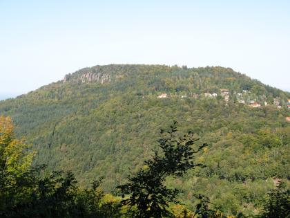 Battert bei Baden-Baden Blick aus großer Entfernung auf einen hohen, bewaldeten Bergrücken. Links oben ragen rötliche Felszinnen aus dem Bergwald, rechts tauchen einzelne Häuser zwischen den Bäumen auf.