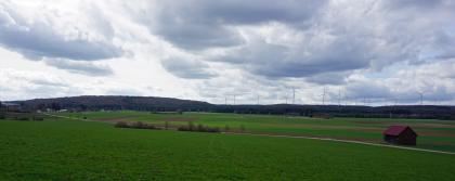 Die Rauhe Wiese bei Böhmenkirch-Heidhöfe – Blick nach Westen über die flache, mit Feuersteinlehm plombierte Karstwanne Blick über eine flachhügelige Wiesenlandschaft, die im Hintergrund von Ackerstreifen unterbrochen sowie von bewaldeten Bergrücken begrenzt wird. Entlang der Bergrücken rechts sind mehrere Windräder zu erkennen. Im Vordergrund verläuft eine schmale Straße.