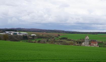 Blick über einen zum Mittelgrund hin abfallenden Wiesenhang. Dahinter, in einer Senke, liegt ein verbuschter Streifen mit Bäumen und Gebäuden. Weiter zum Hintergrund hin folgen flache Wiesen und Äcker sowie ein langer bewaldeter Bergrücken.