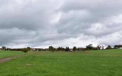In den Mulden der Hochflächenränder, wie hier bei Ebhausen-Ebershardt, gibt es örtlich Quellaustritte und grundwasserbeeinflusste Böden Blick auf eine leicht wellige, mit Gras bewachsene Hochfläche. Links führt ein Feldweg auf im Hintergrund stehende Bäume und Häuser zu.