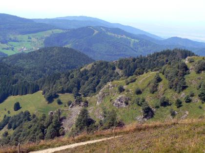 Blick vom Belchen (1414 m ü. NHN) nach Südwesten zum Blauen und rechts hinten in die Oberrheinebene Blick aus großer Höhe auf einen felsigen, nur teilweise bewaldeten Berghang, der sich nach rechts hinaufzieht. Dahinter sind weitere Berge zu sehen, links oben auch eine Hochfläche. Rechts oben ist im Dunst eine weite Ebene erkennbar.