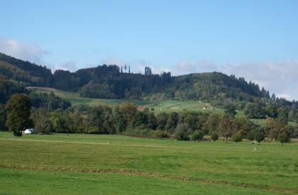 Weitenauer Bergland – Vorne die Aue der Kleinen Wiese, dahinter die Kreismülldeponie Scheinberg Hinter einer Wiesenlandschaft im Vordergrund sowie einem Baumgürtel erhebt sich ein nach links ansteigender, auf der Kuppe bewaldeter Berg.