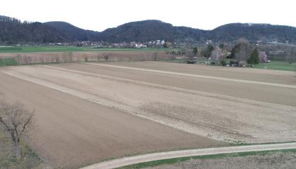 Hochrheintal bei Rheinfelden-Degerfelden – Blick zum Dinkelberg Blick auf weite flache Ackerflächen, diagonal von links oben nach rechts unten gefurcht. Im Hintergrund Wiesen, Bäume, einige Häuser sowie mehrere bewaldete Berge. Im Berg rechts sind Straßentunnel erkennbar.